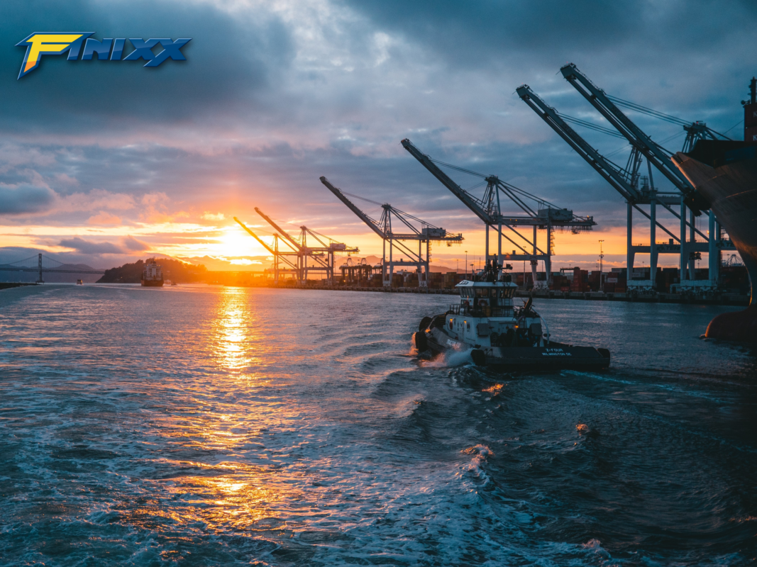 Sunset view of a shipping port with cranes and a tugboat navigating the water, representing Finixx global transportation and logistics.